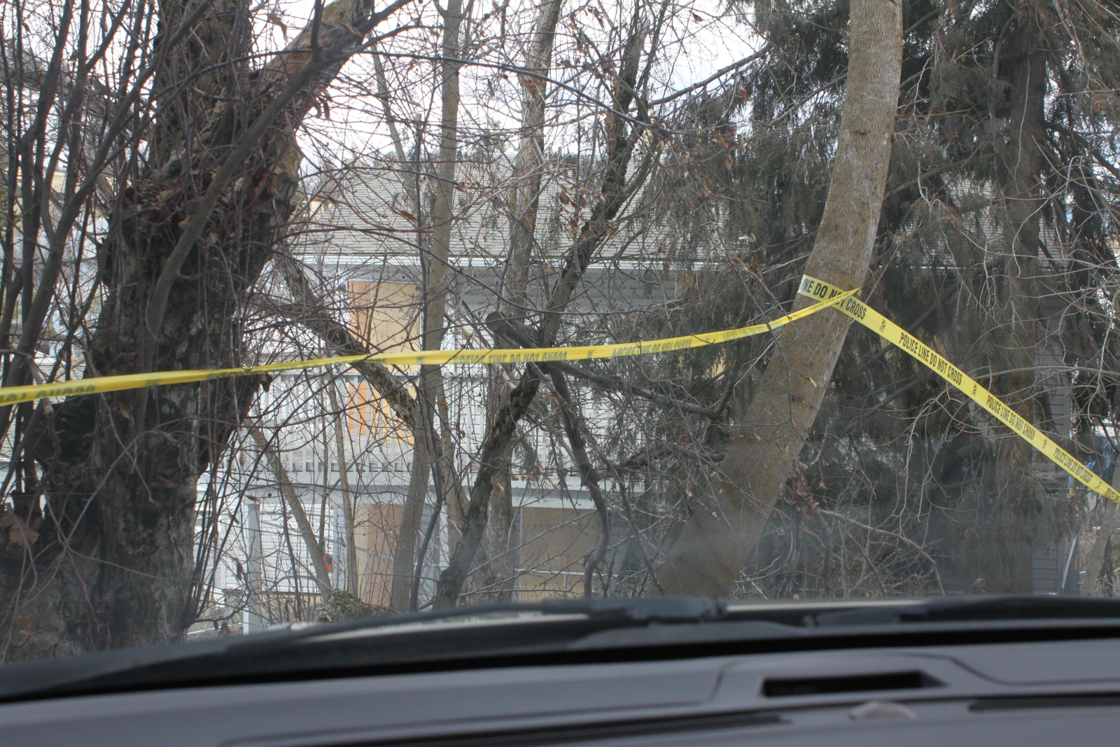 View through a vehicle windshield toward bare trees and a light-colored house, with yellow caution tape tied between trees and a dashboard below.