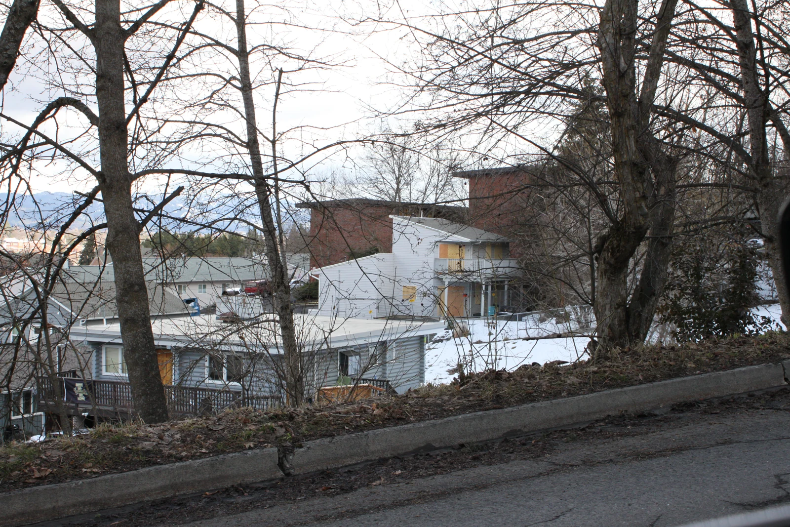 Wide roadside view of a light-colored multi-level house below bare trees, with snow on the slope and boarded windows.