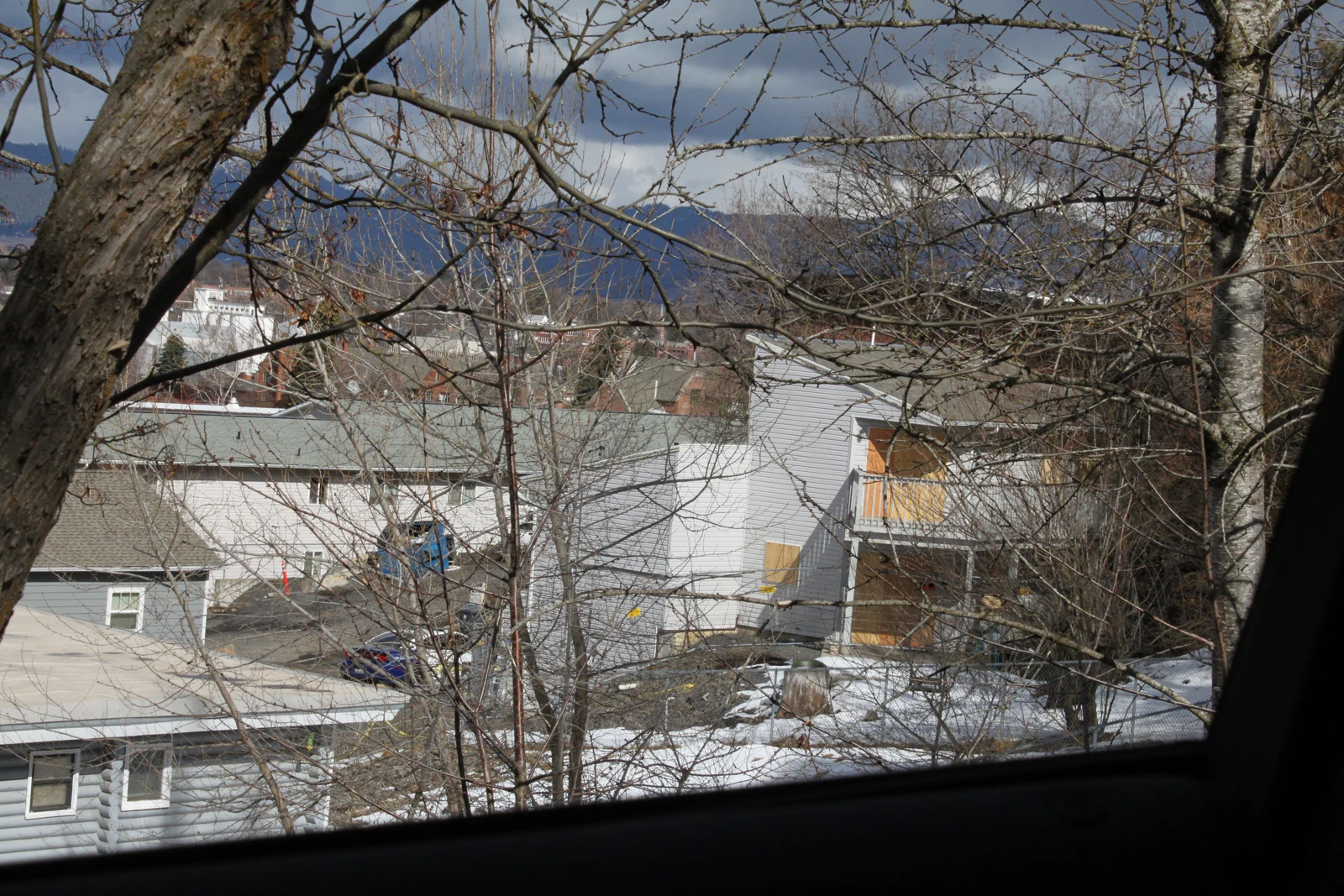 View from a vehicle toward a light-colored house behind bare branches, with a dark window edge across the bottom.