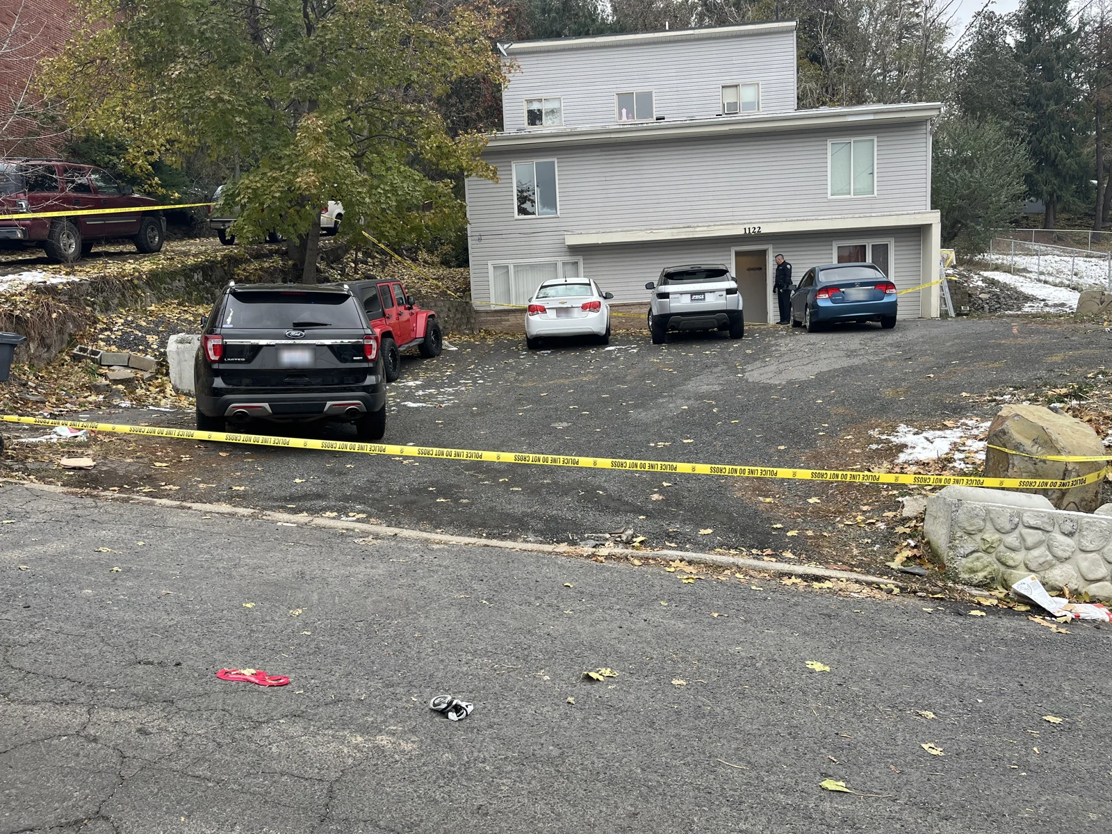 Front of a gray multi-level house with several parked vehicles, yellow caution tape across the driveway and scattered objects in the road.