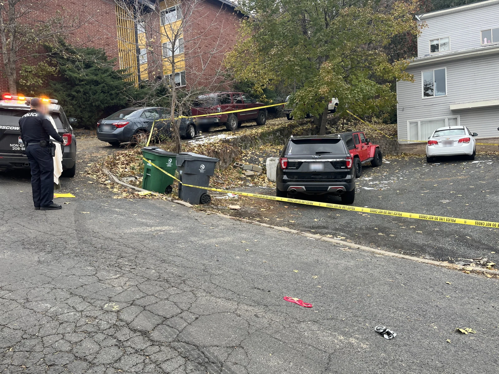 Street and driveway beside a gray house, with parked vehicles, trash bins, yellow police tape and a uniformed person near a vehicle.