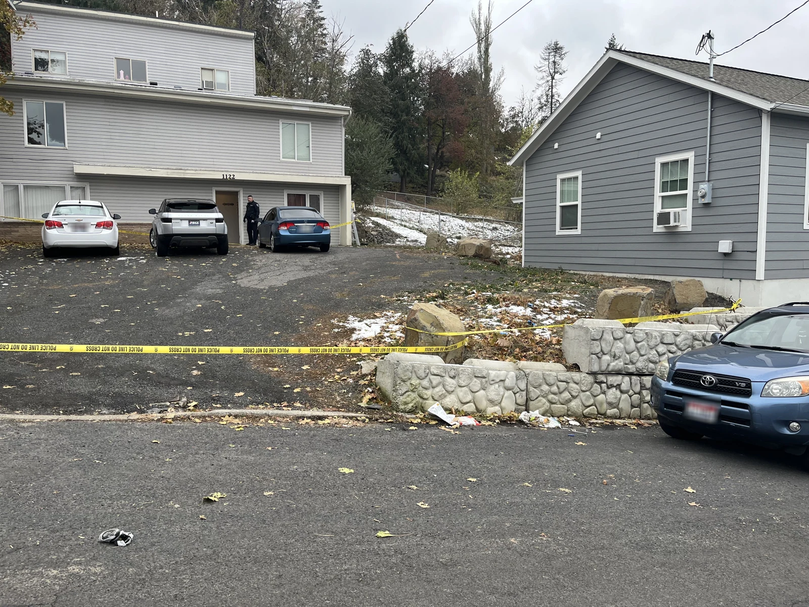 Driveway between gray buildings with several parked vehicles, yellow police tape and a low stone retaining wall.