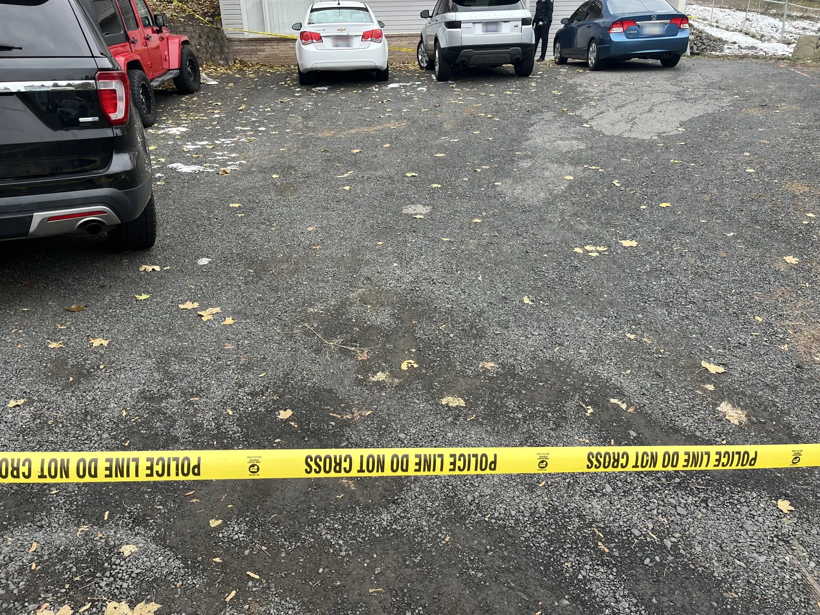 Asphalt driveway with fallen leaves, parked vehicles and yellow Police Line Do Not Cross tape across the foreground.