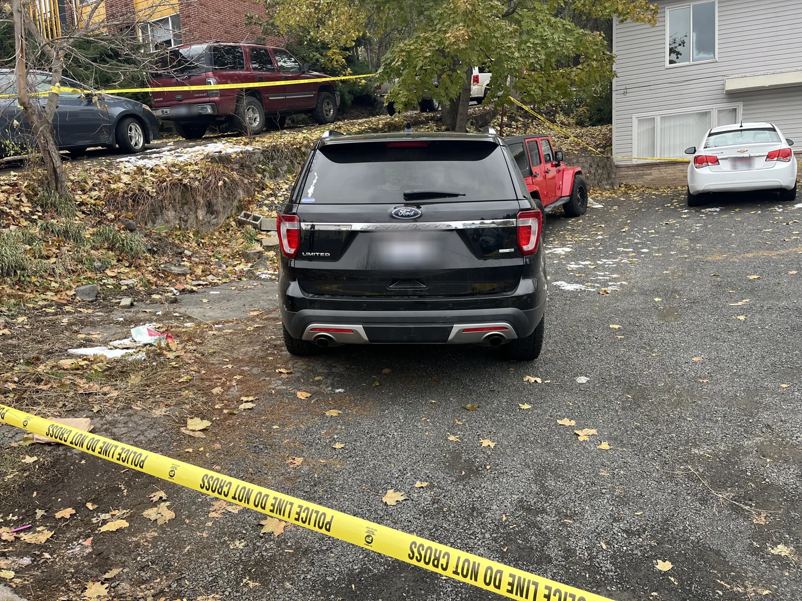 Rear of a black SUV in a driveway, with a red Jeep and white sedan ahead and yellow police tape across the foreground.
