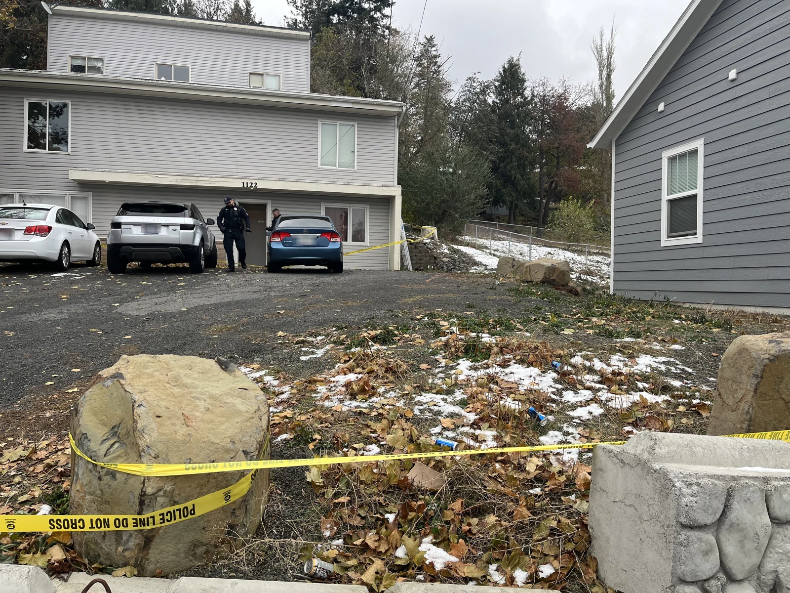 Gray house entrance with three parked vehicles and a standing person, seen across yellow tape, rocks and patches of snow.
