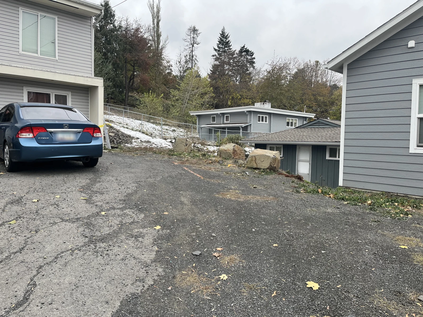 Sloping asphalt driveway between gray buildings, with a blue sedan, fallen leaves, rocks and snow along the side.