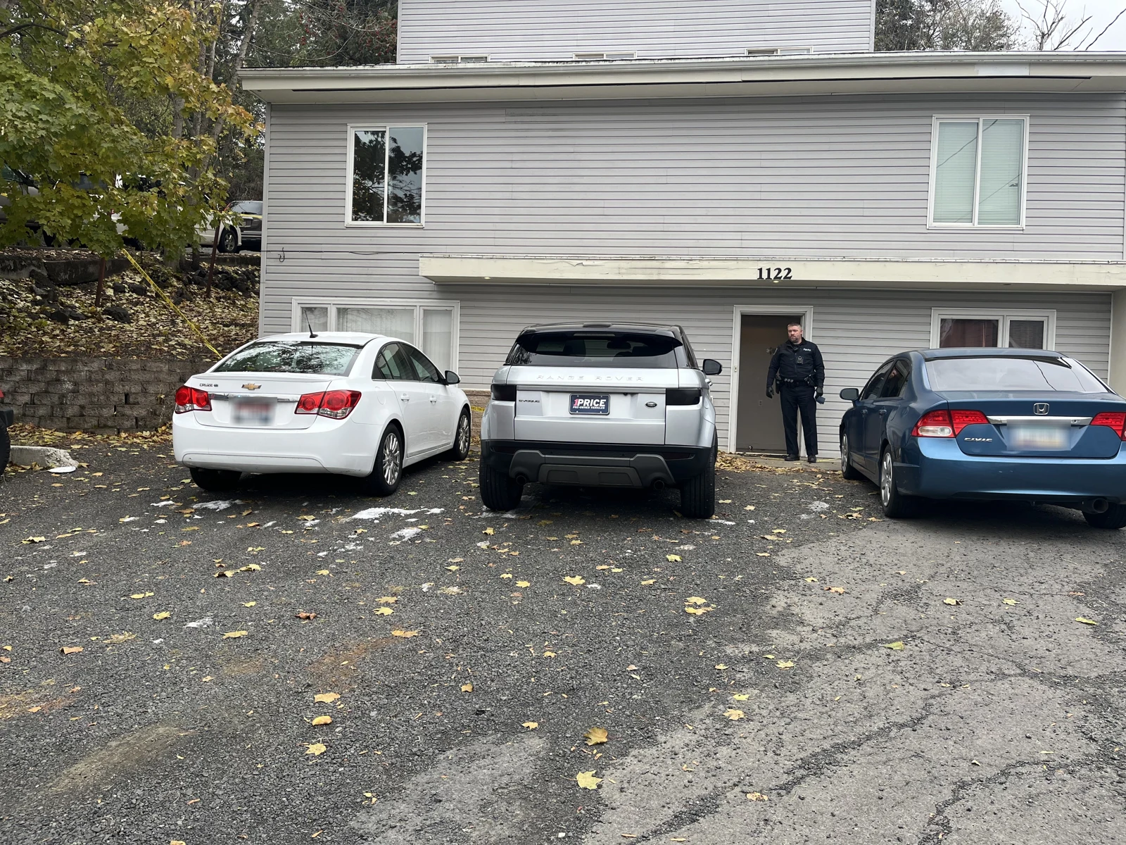 Front entrance of a gray house marked 1122, with a white sedan, silver SUV, blue sedan and a standing uniformed person.