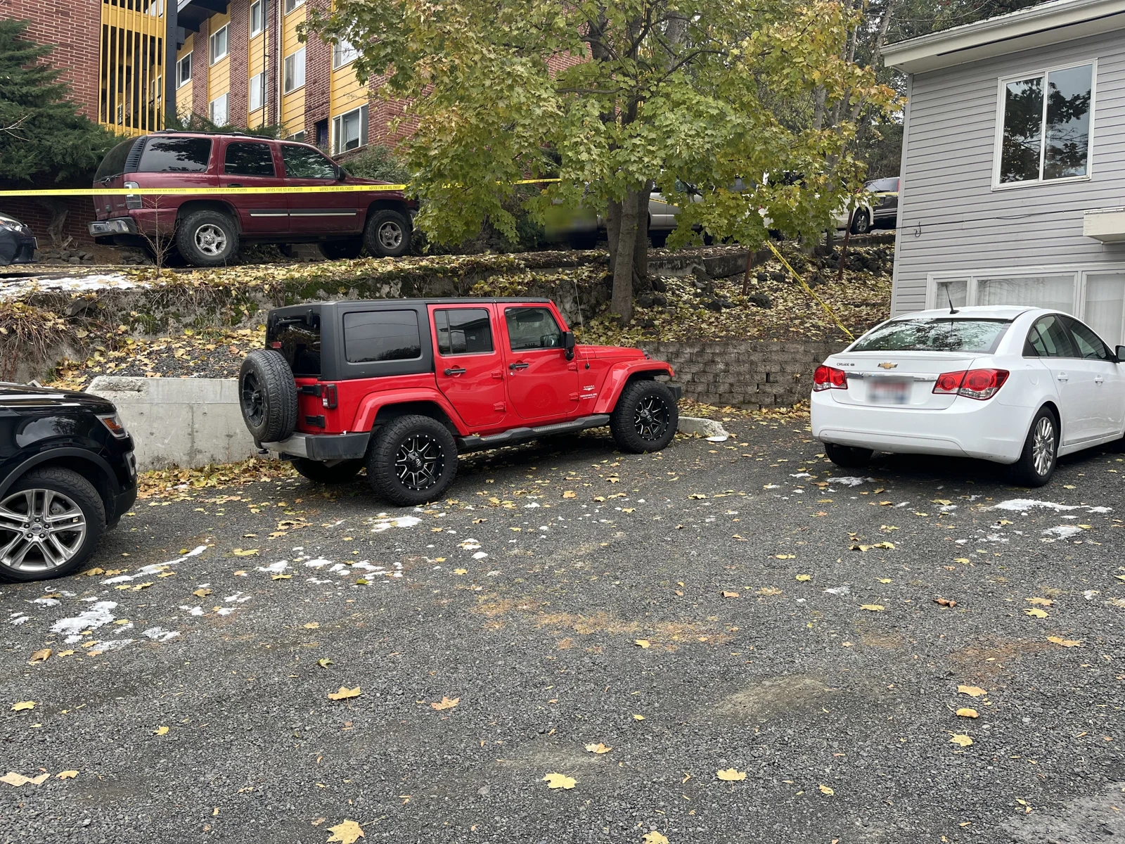Red Jeep and white sedan in a driveway, with a maroon vehicle on the raised ground behind a retaining wall and yellow tape.