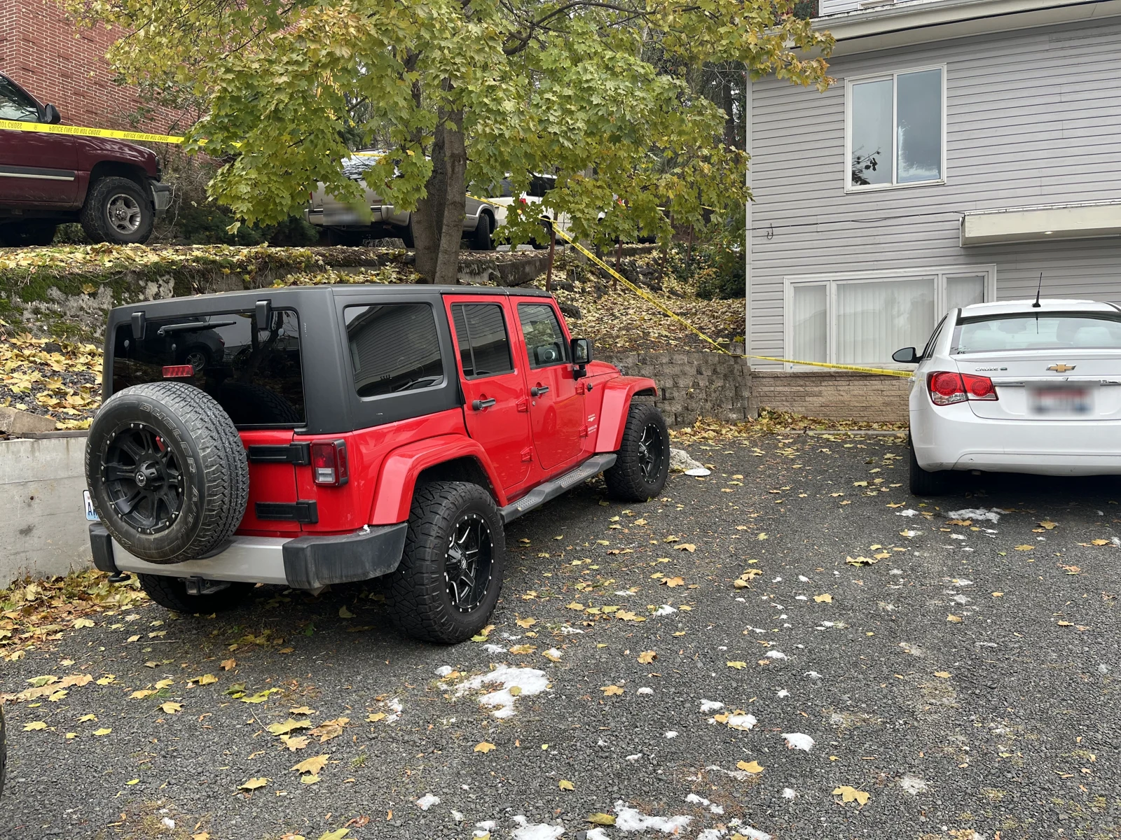 Rear-side view of a red Jeep with a black roof and spare tire, parked beside a white sedan and patches of snow.