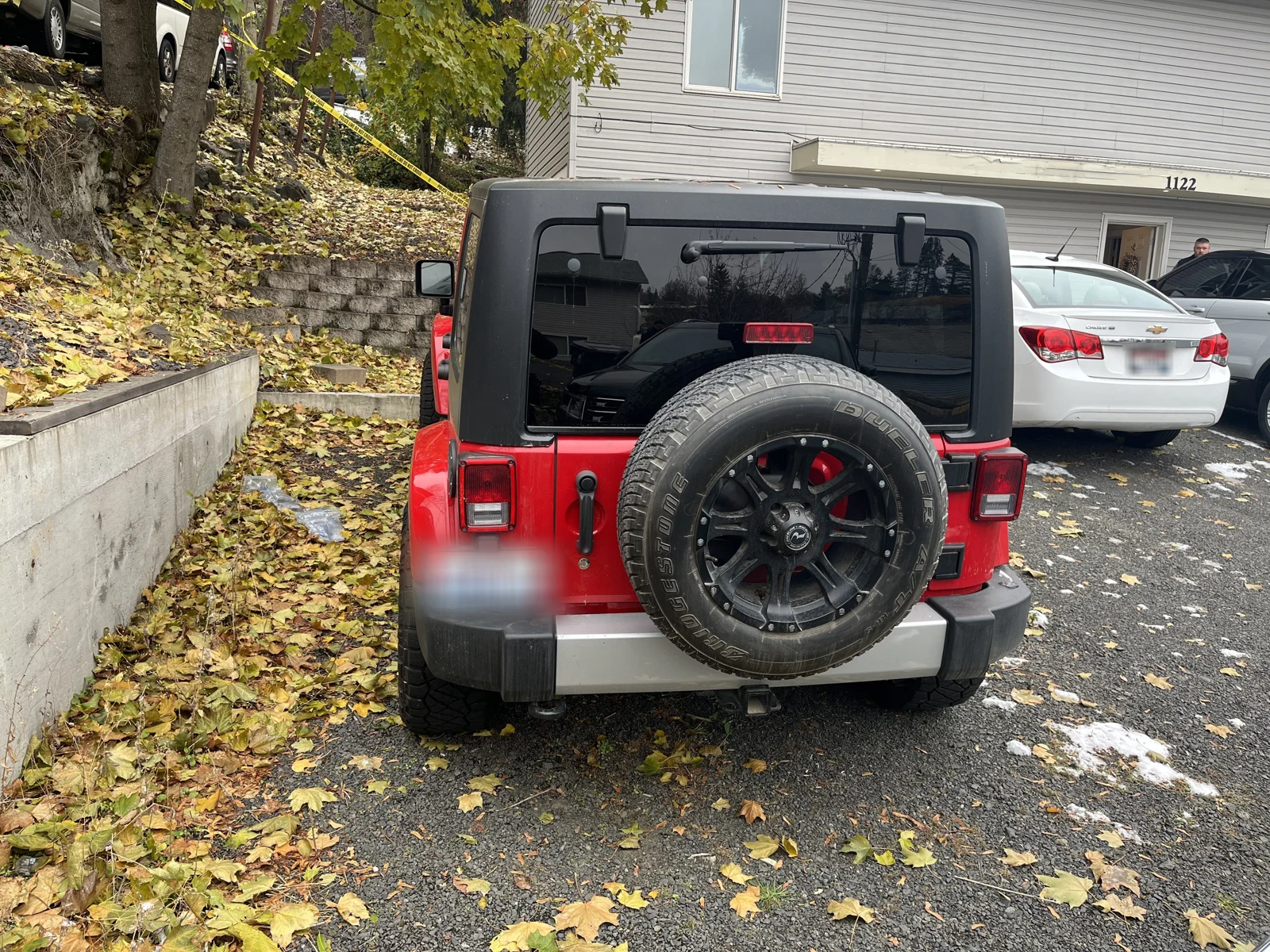 Rear of a red Jeep with a spare tire and blurred license plate, parked beside a retaining wall and fallen leaves.
