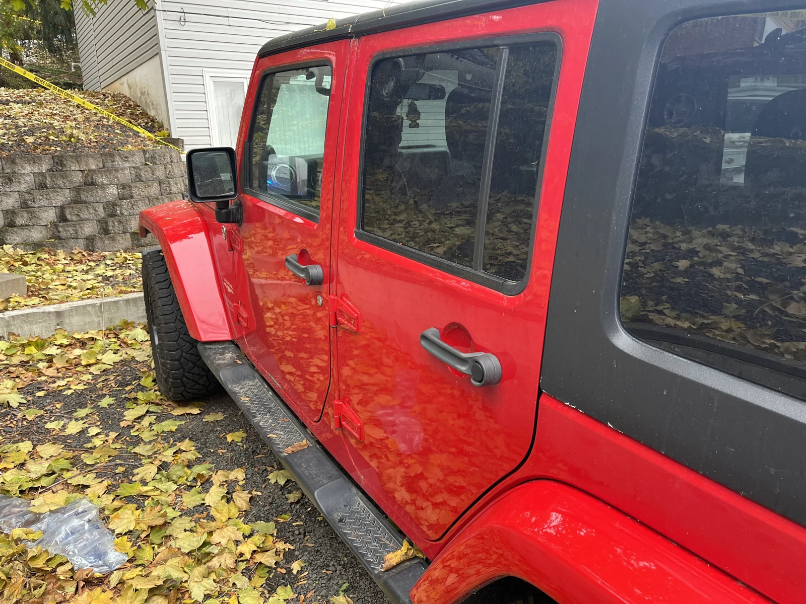 Side of a red four-door Jeep with black roof and reflective windows, parked beside fallen leaves and yellow tape.