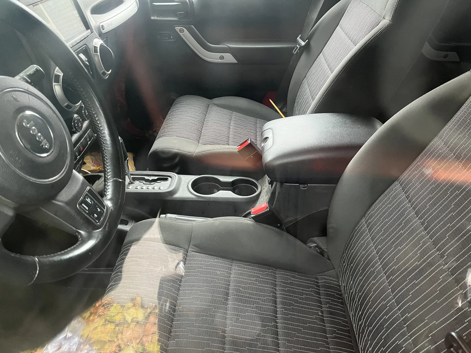 Front seats, Jeep steering wheel, gear selector and two cupholders inside a vehicle, viewed through a window.