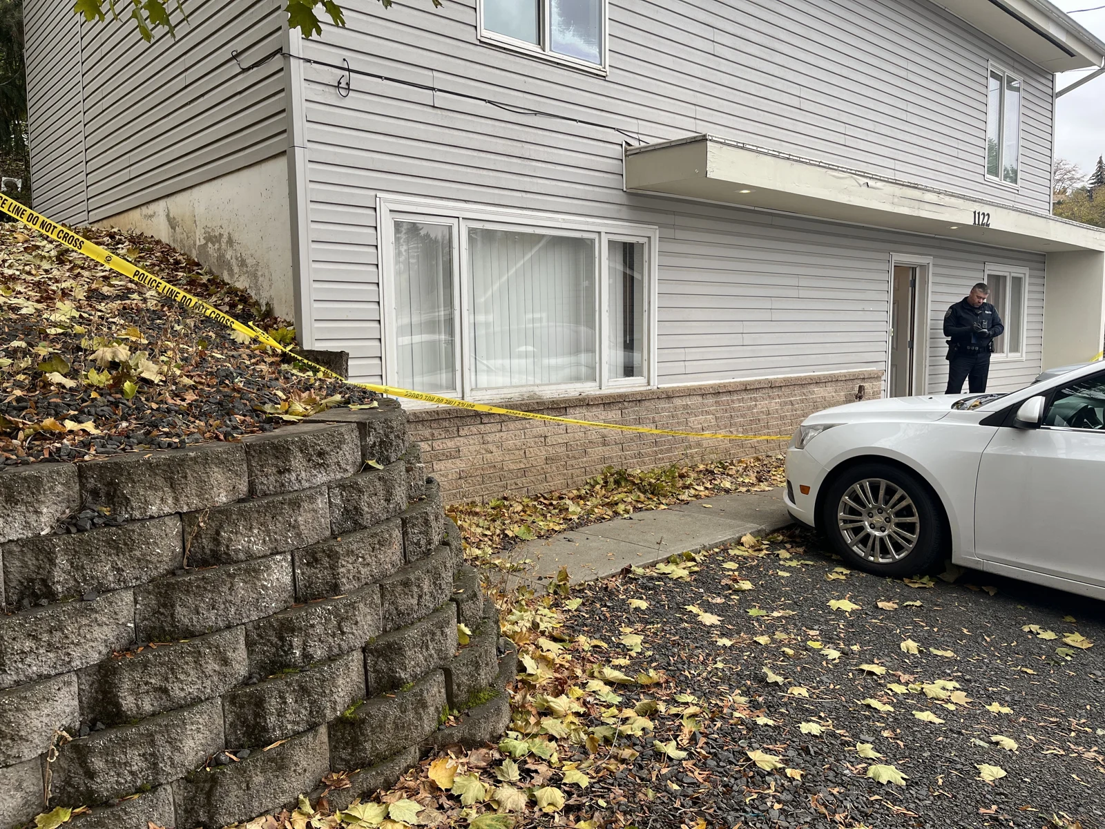 Gray house exterior, retaining wall and white sedan, with yellow tape and a uniformed person by the doorway.