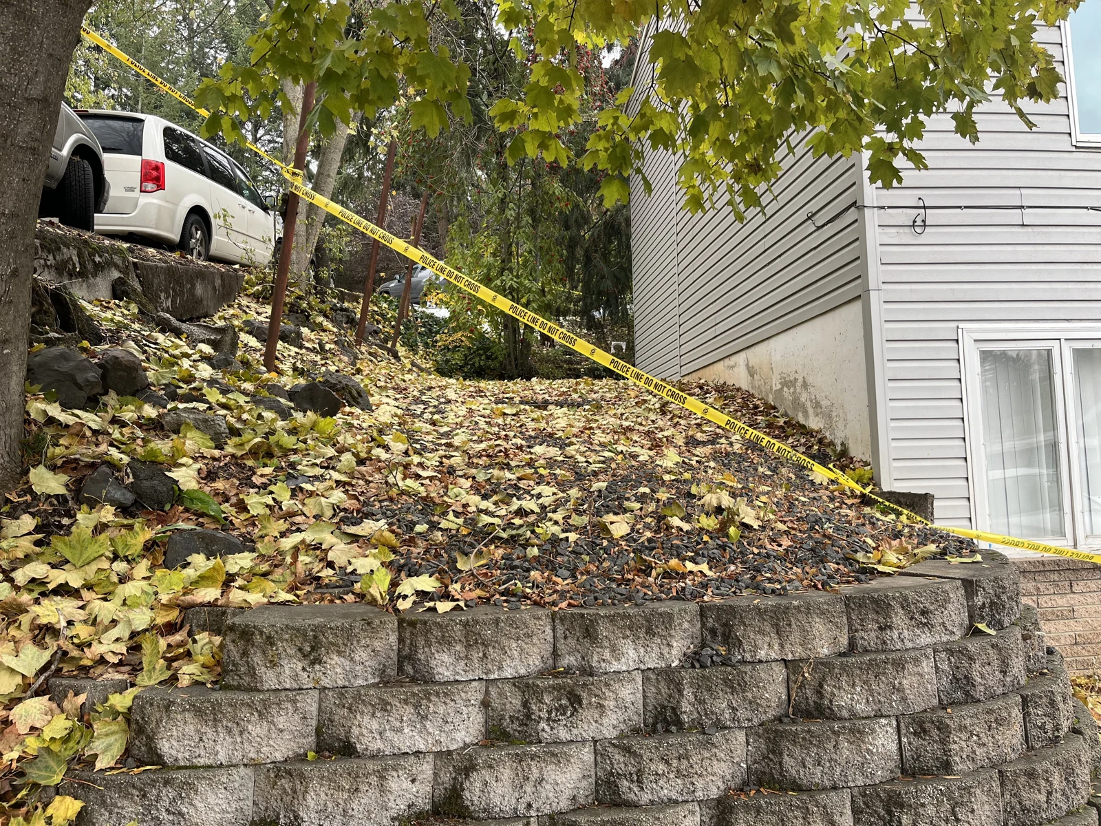 Leaf-covered slope and concrete retaining wall beside a gray house, with a tree, yellow tape and a white SUV above.