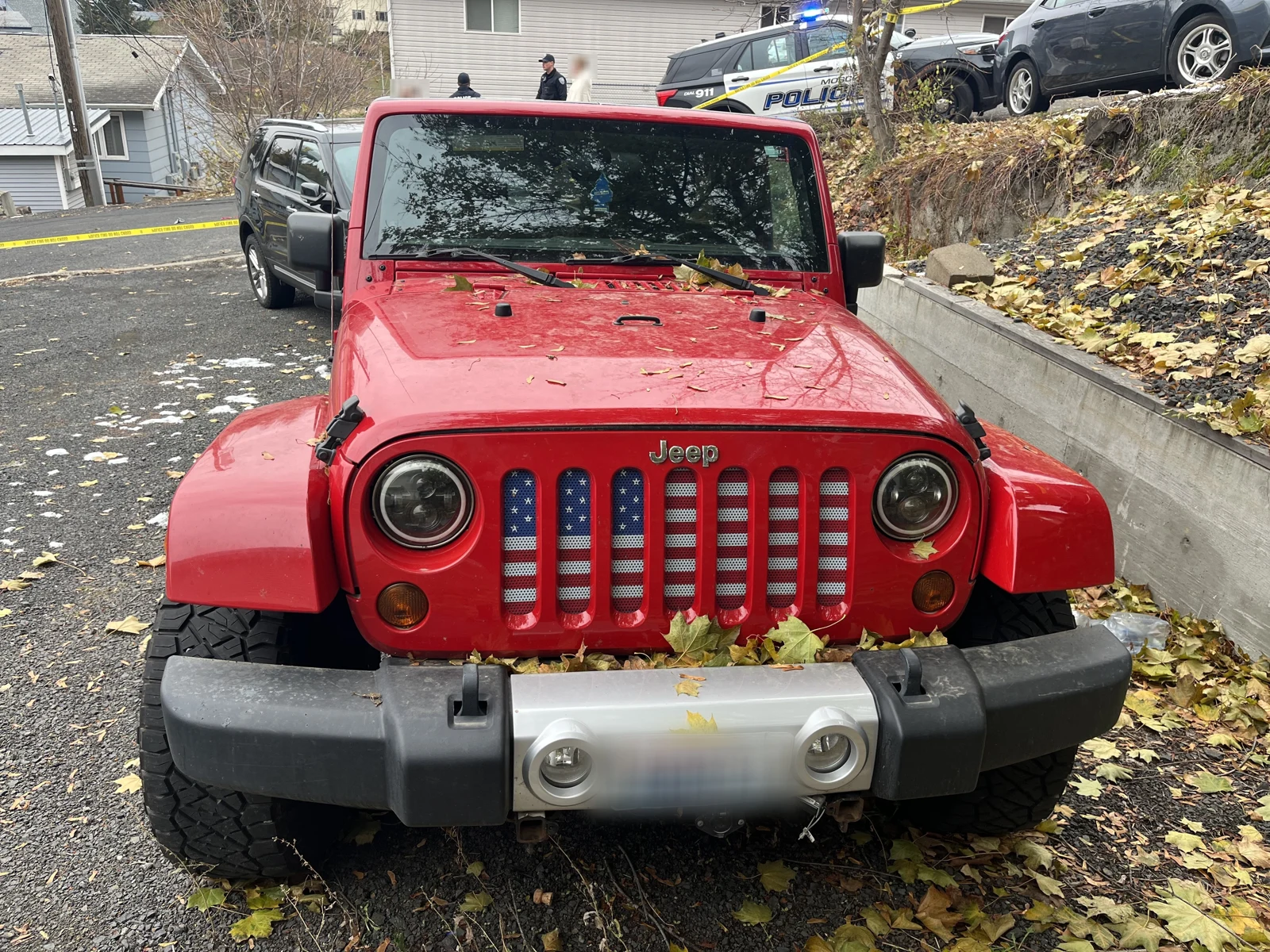 Front of a red Jeep with a flag-pattern grille, fallen leaves on the hood and a blurred license plate; police vehicles are uphill.