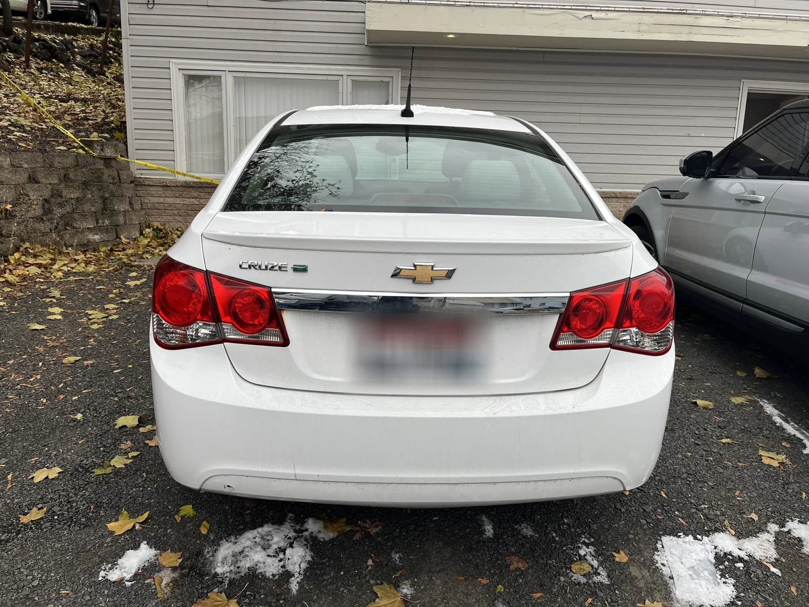 Rear of a white Chevrolet Cruze with a blurred plate, parked beside a silver SUV and patches of snow.