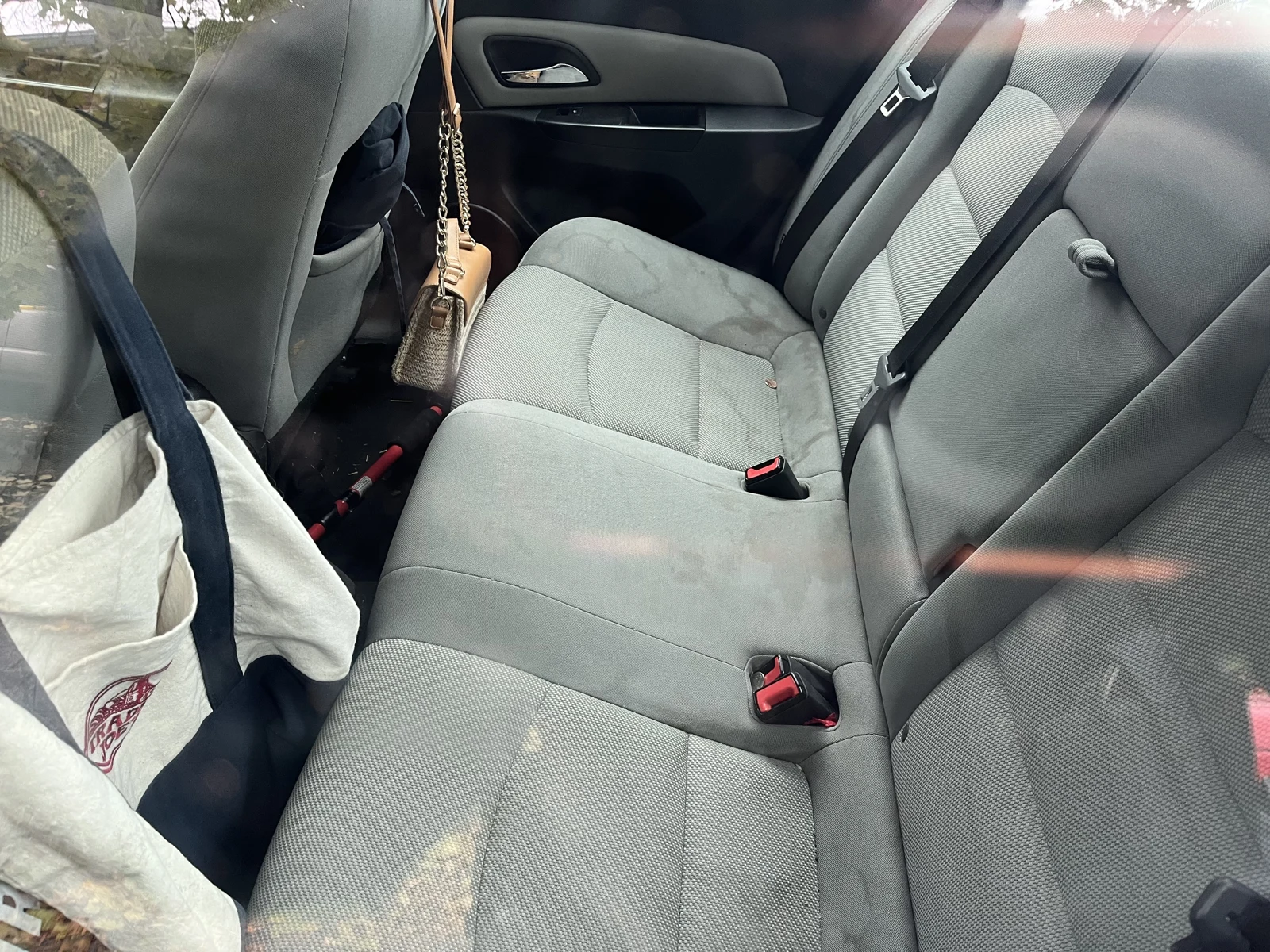 Light-gray rear bench seat with seat belts, a hanging handbag and pale fabric in the footwell, seen through reflective glass.