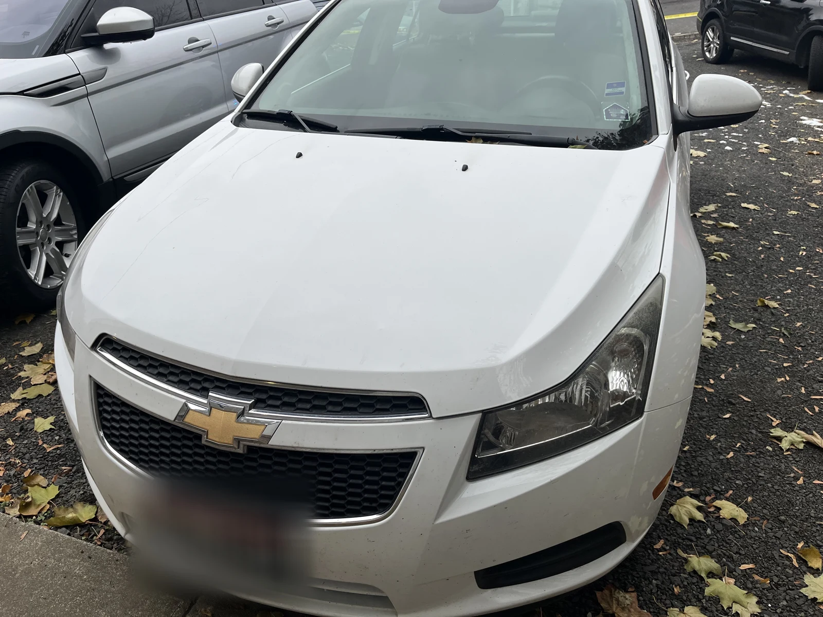 Front of a white Chevrolet sedan with a gold bowtie emblem, blurred plate and fallen leaves around it.