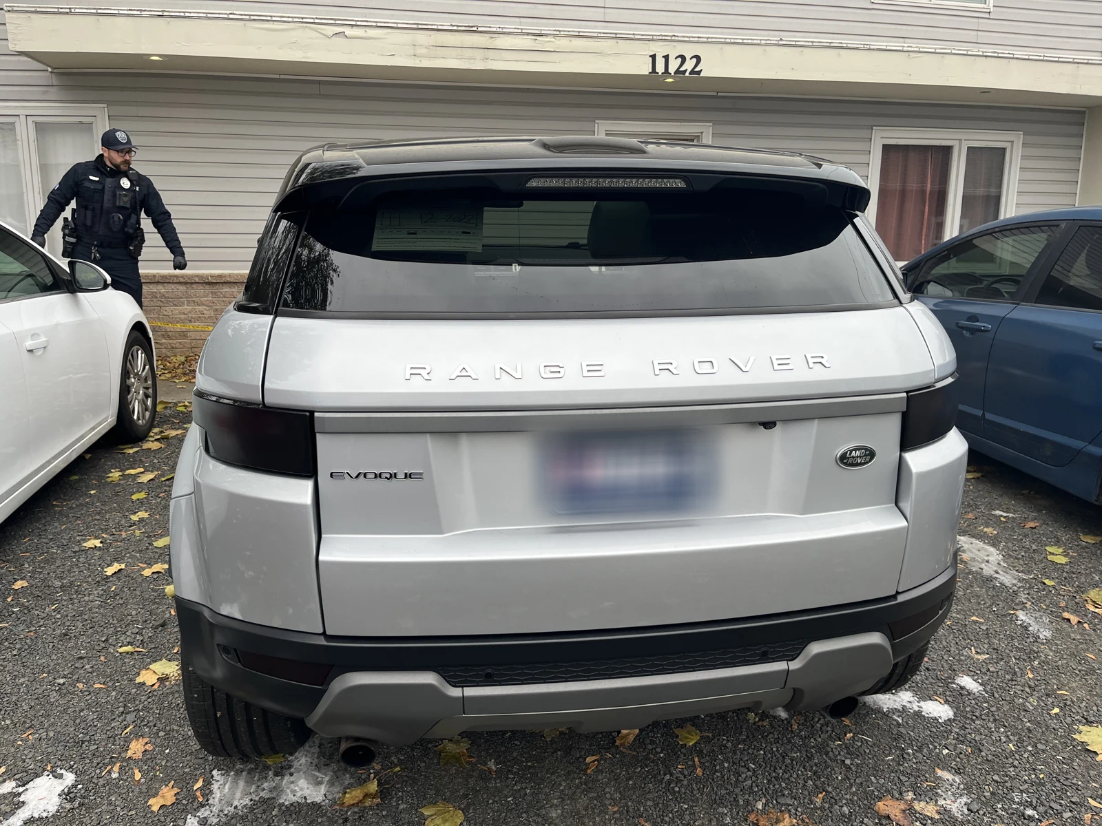 Rear of a silver Range Rover Evoque with a blurred plate, parked outside a house marked 1122 near a uniformed person.