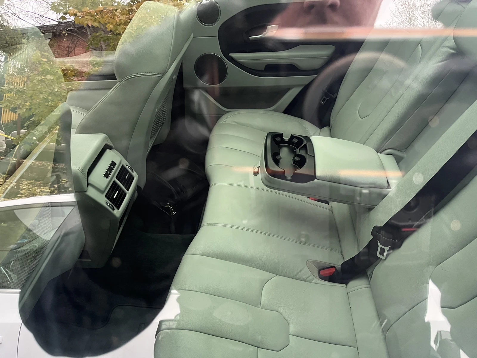 Light-colored leather rear bench seat with a folded-down center armrest and cupholders, viewed through reflective vehicle glass.