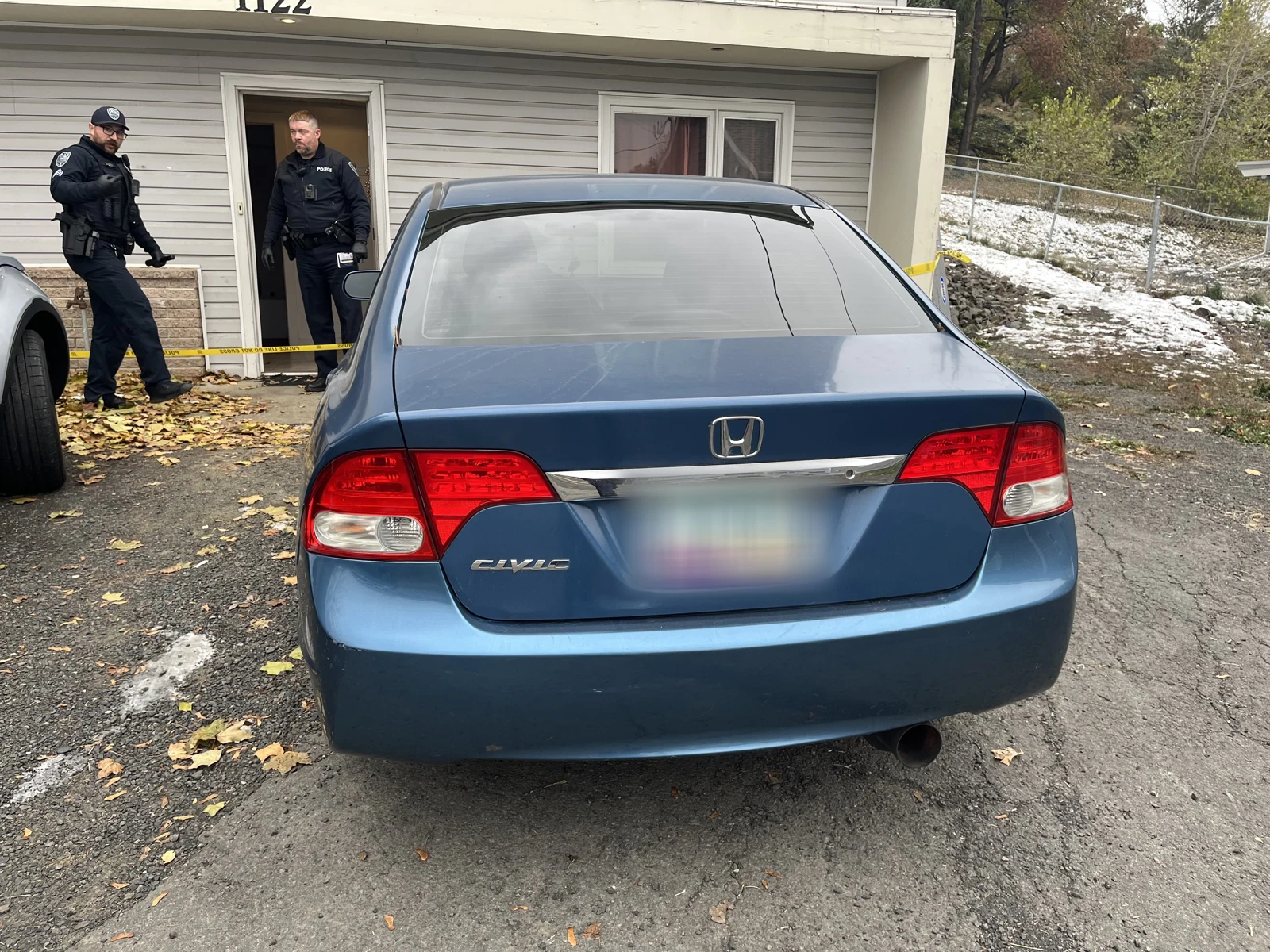 Rear of a blue Honda Civic with a blurred plate, parked outside a gray house while two uniformed people stand near the entrance.