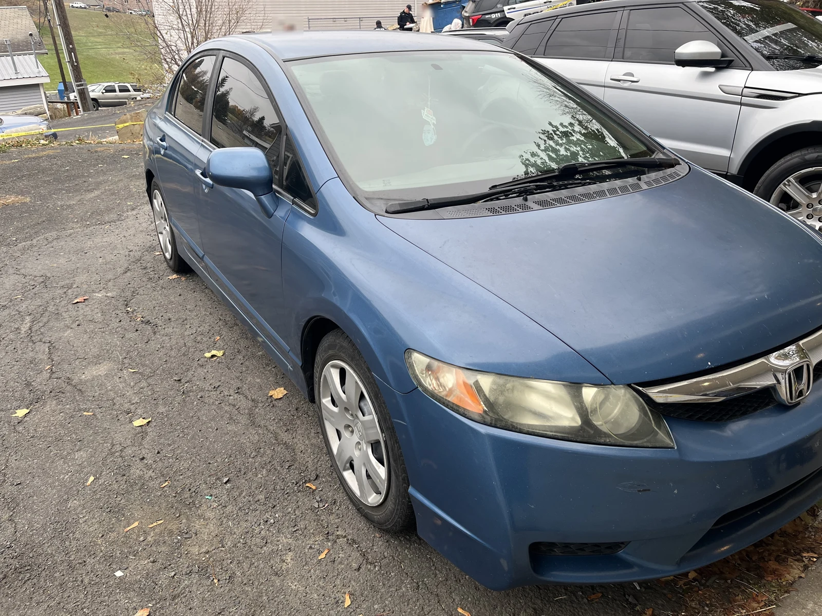 Front and side of a blue Honda sedan parked beside a silver SUV on a leaf-strewn driveway.