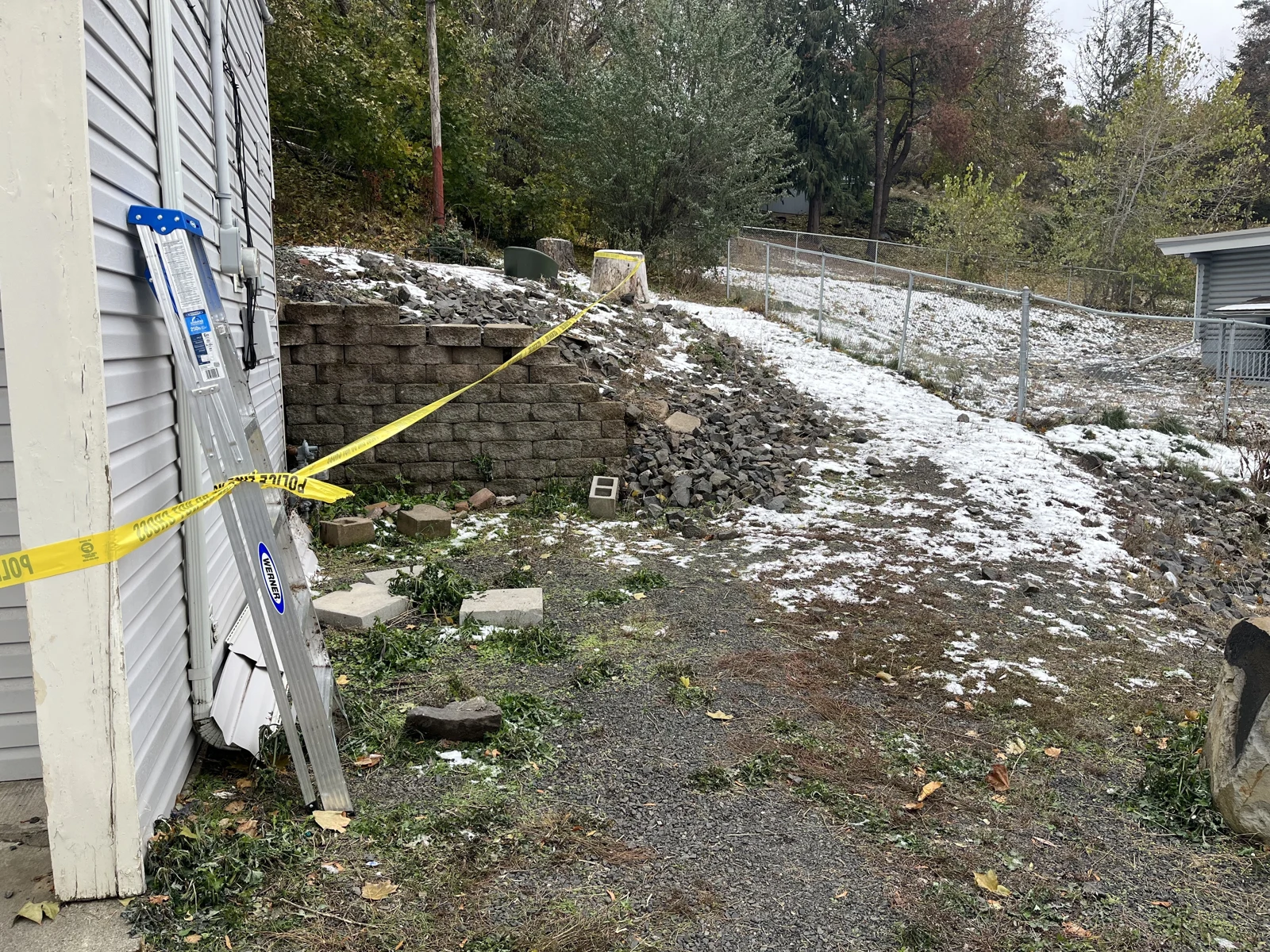 Narrow ground area beside gray siding with a ladder, concrete blocks, retaining wall, chain-link fence and patches of snow.