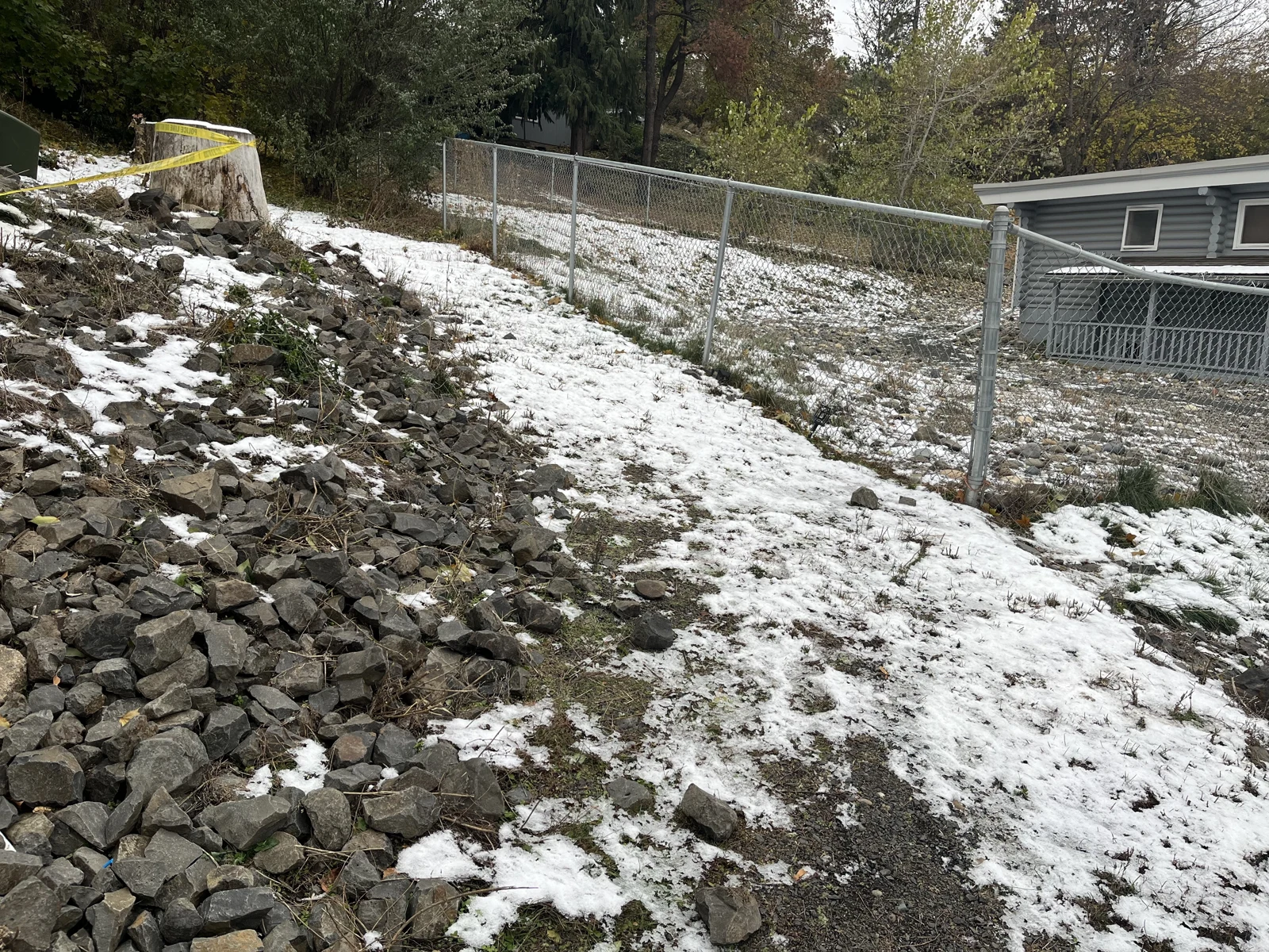 Snow-covered slope with loose rocks, a tree stump wrapped in yellow tape and a chain-link fence.