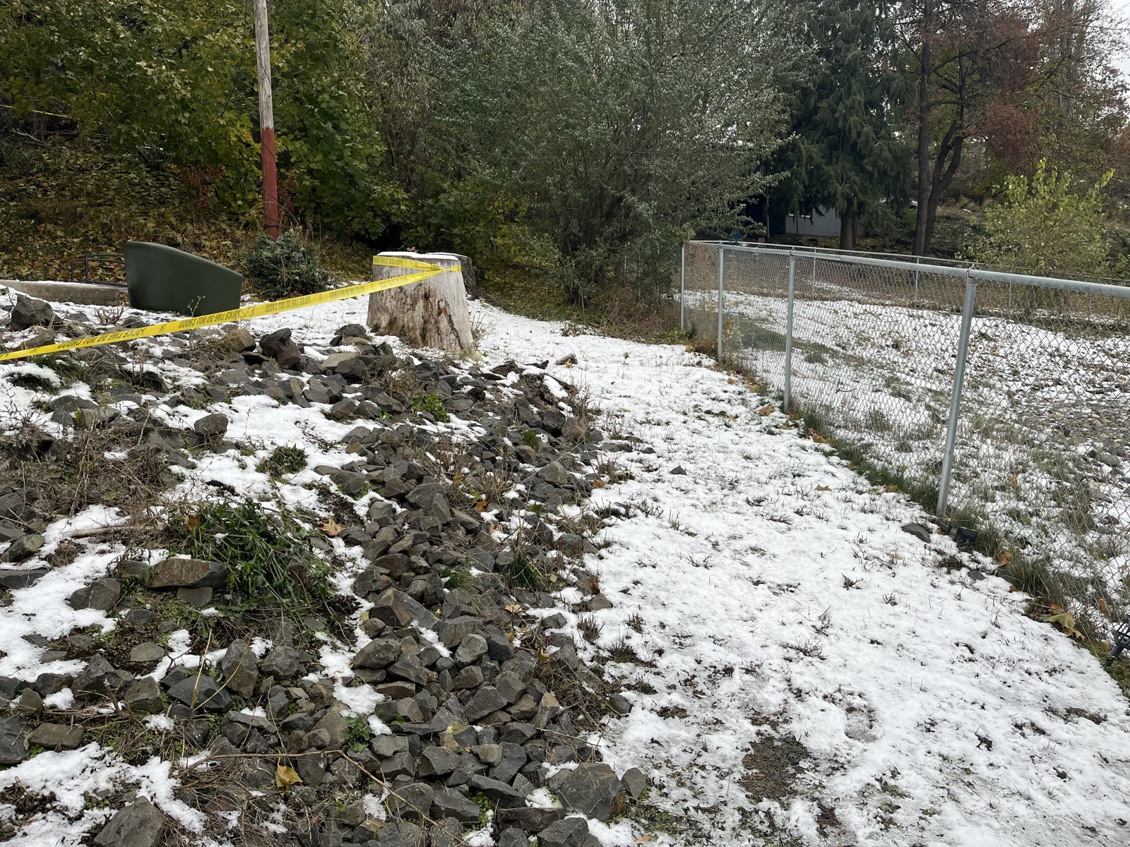 Wider view of a rocky, snow-covered slope beside a chain-link fence, with trees and yellow caution tape.