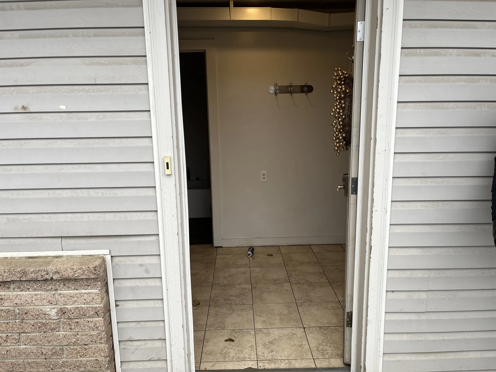 Open exterior door in gray siding, looking into a tiled entry hall with wall hooks and small objects on the floor.