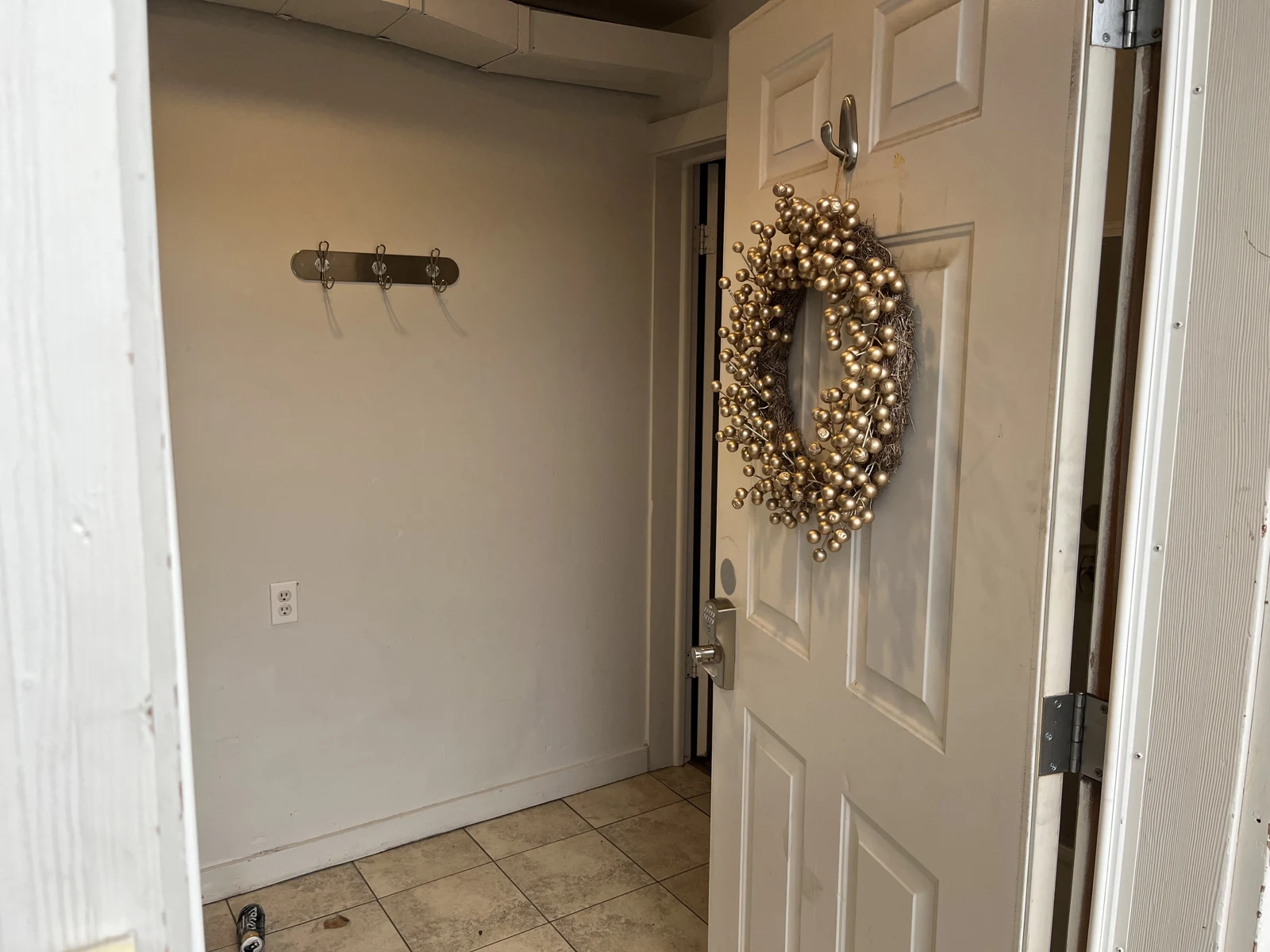 Open white front door with a gold decorative wreath, beside a tiled entry hall and wall-mounted coat hooks.