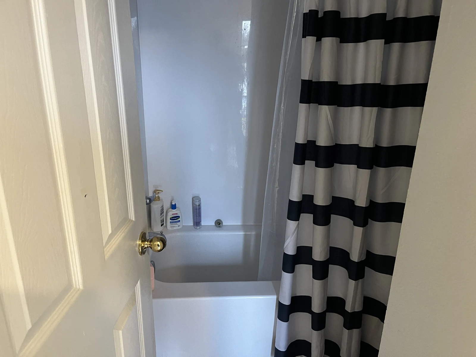 Bathroom doorway showing a bathtub, toiletries and a black-and-white horizontally striped shower curtain.