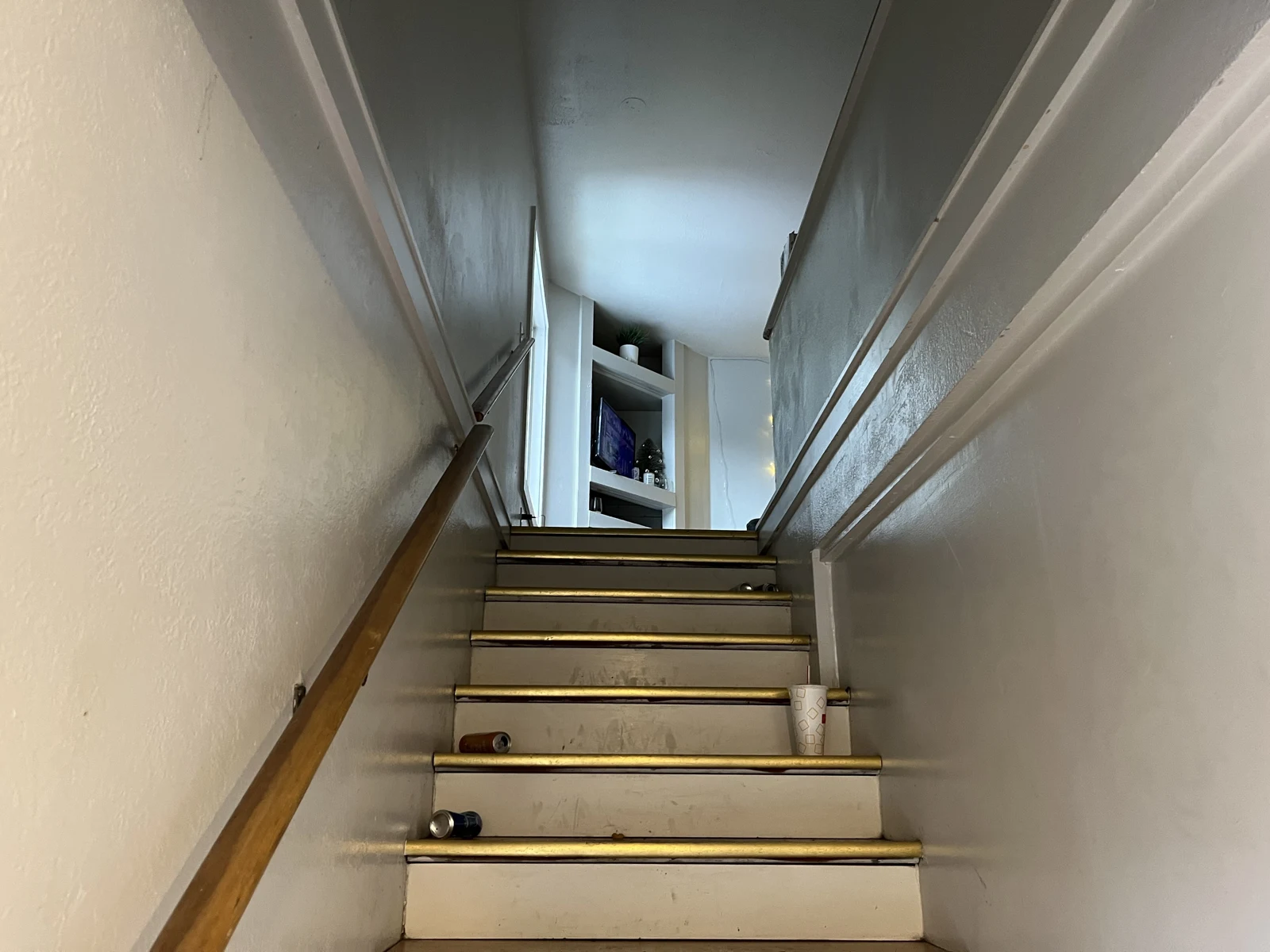 Upward view of a narrow wooden staircase with handrails, a cup and small objects on the steps, leading toward a television area.