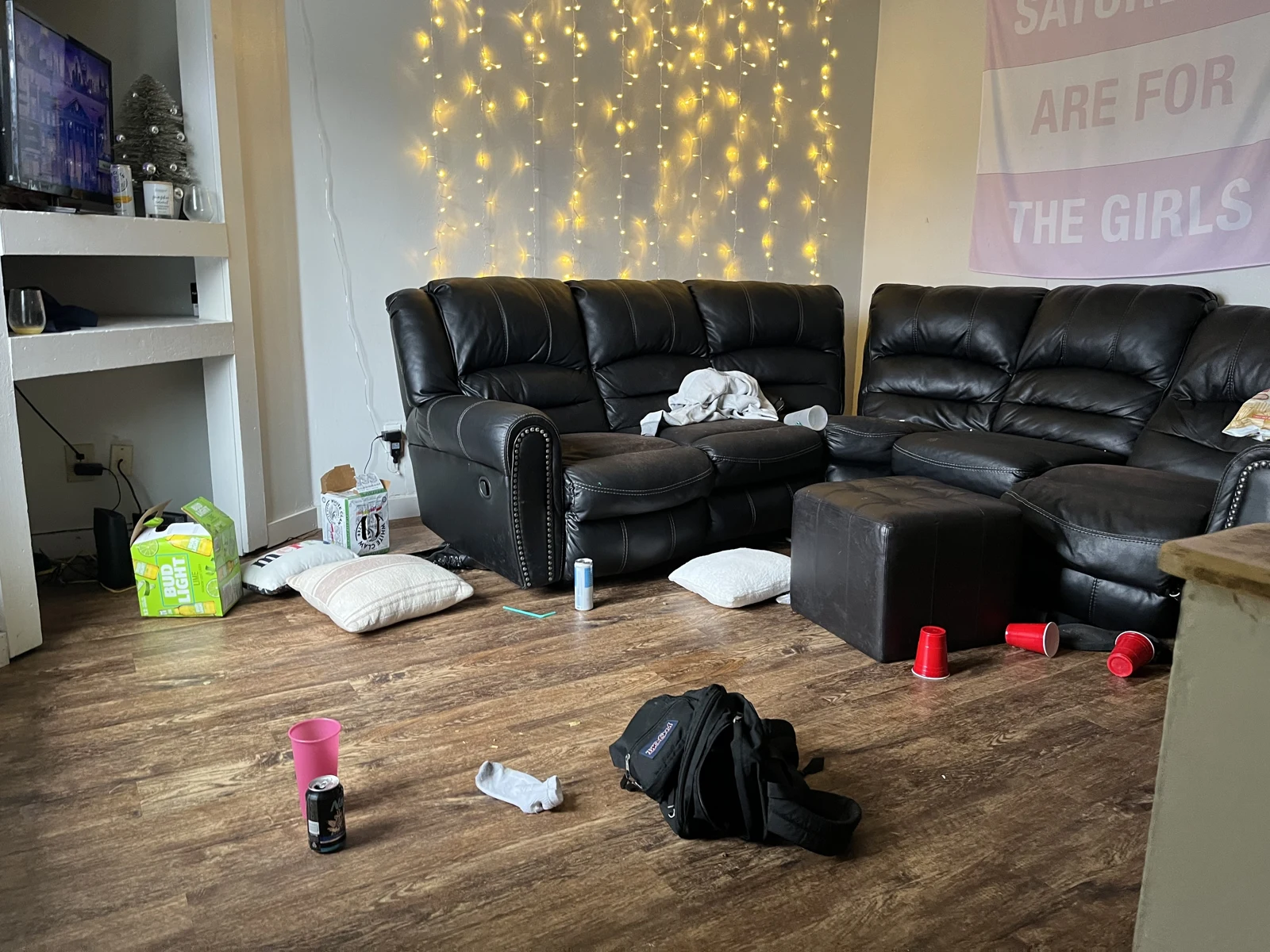 Living room with black leather sectional seating, hanging string lights, red cups, floor cushions, a backpack and a television on shelving.