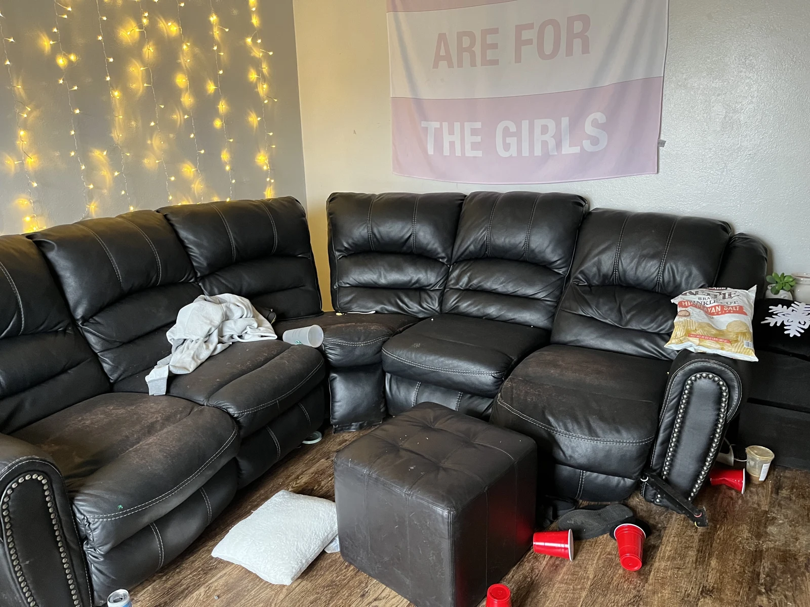 Black leather sectional sofa and square ottoman beneath string lights and a wall banner, with cushions, cups and clothing scattered nearby.