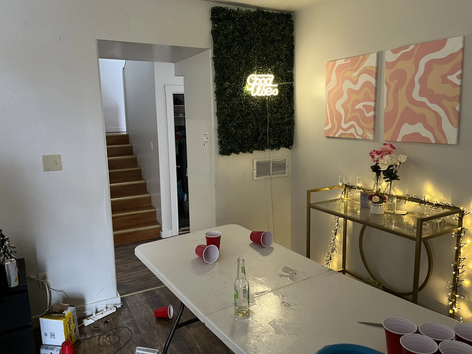 Living-room corner with a white folding table, red cups and a bottle, a gold console table, flowers, decorative lights and stairs through a doorway.