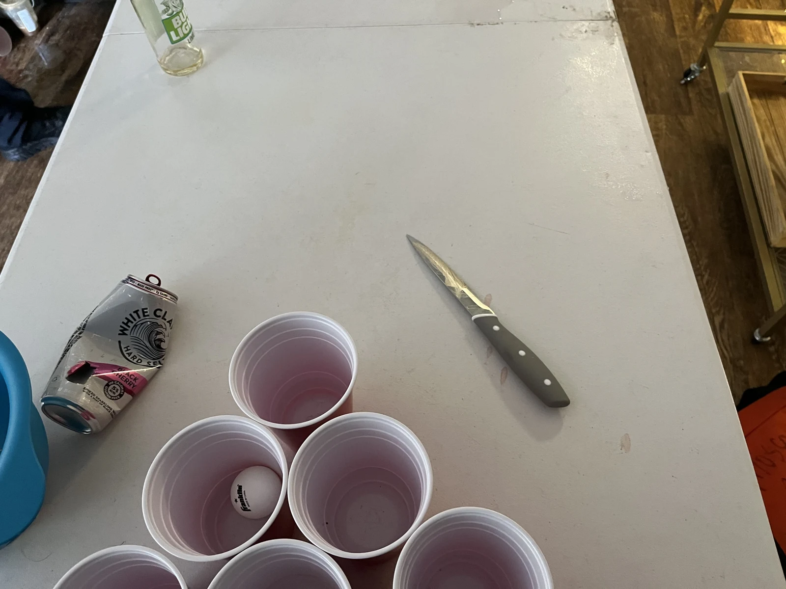 Close view of a white cup-game folding table with clustered red plastic cups, a small white ball in a cup, a handled knife, a can and a bottle.