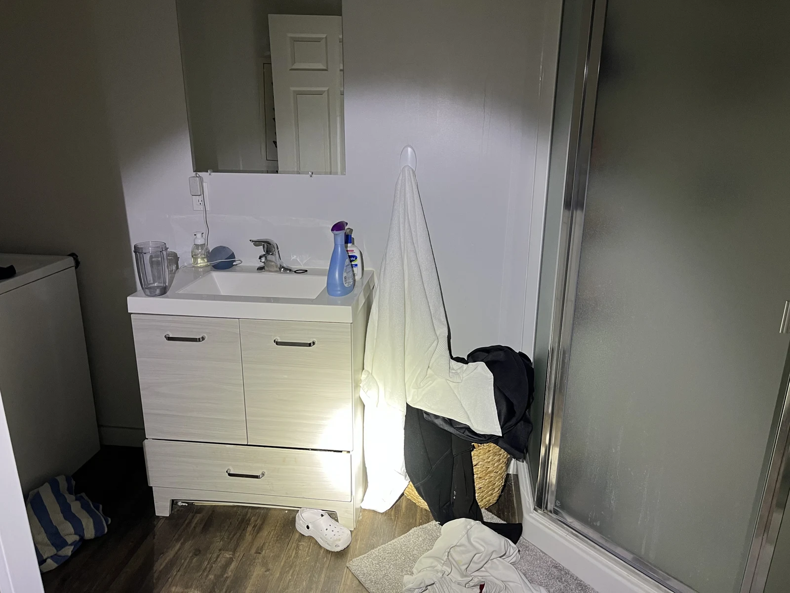 Bathroom vanity with sink, mirror and toiletries beside a frosted shower, towel, basket and a white clog on the floor.