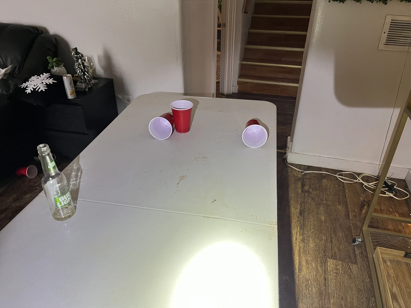 White folding table with red cups and a glass bottle, with a sofa to the left and stairs through a doorway beyond.