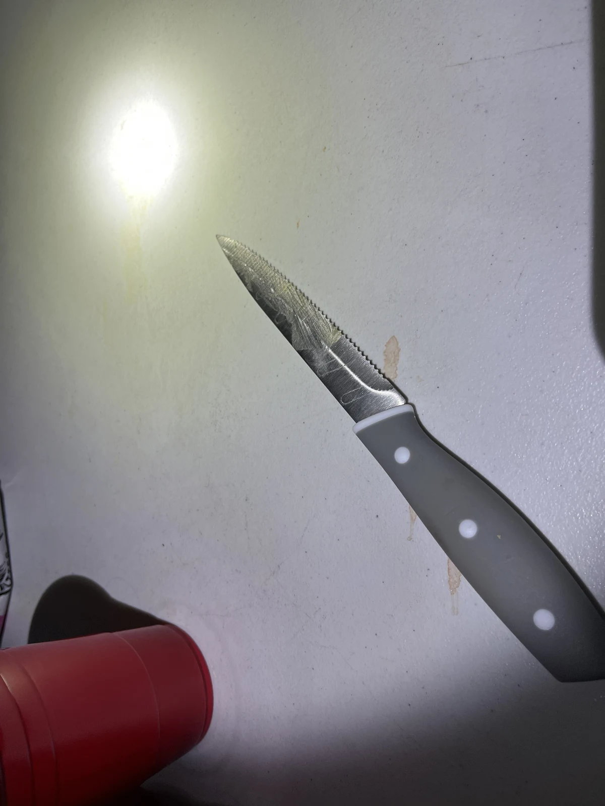 Close photograph of a small pointed knife with a dark three-rivet handle on a white tabletop, beside a red cup and flashlight glare.