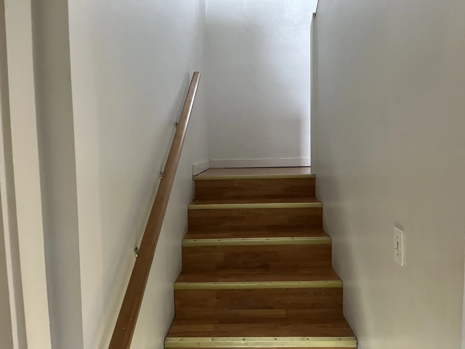 Narrow wooden staircase with white walls and a wooden handrail, leading upward around a corner.