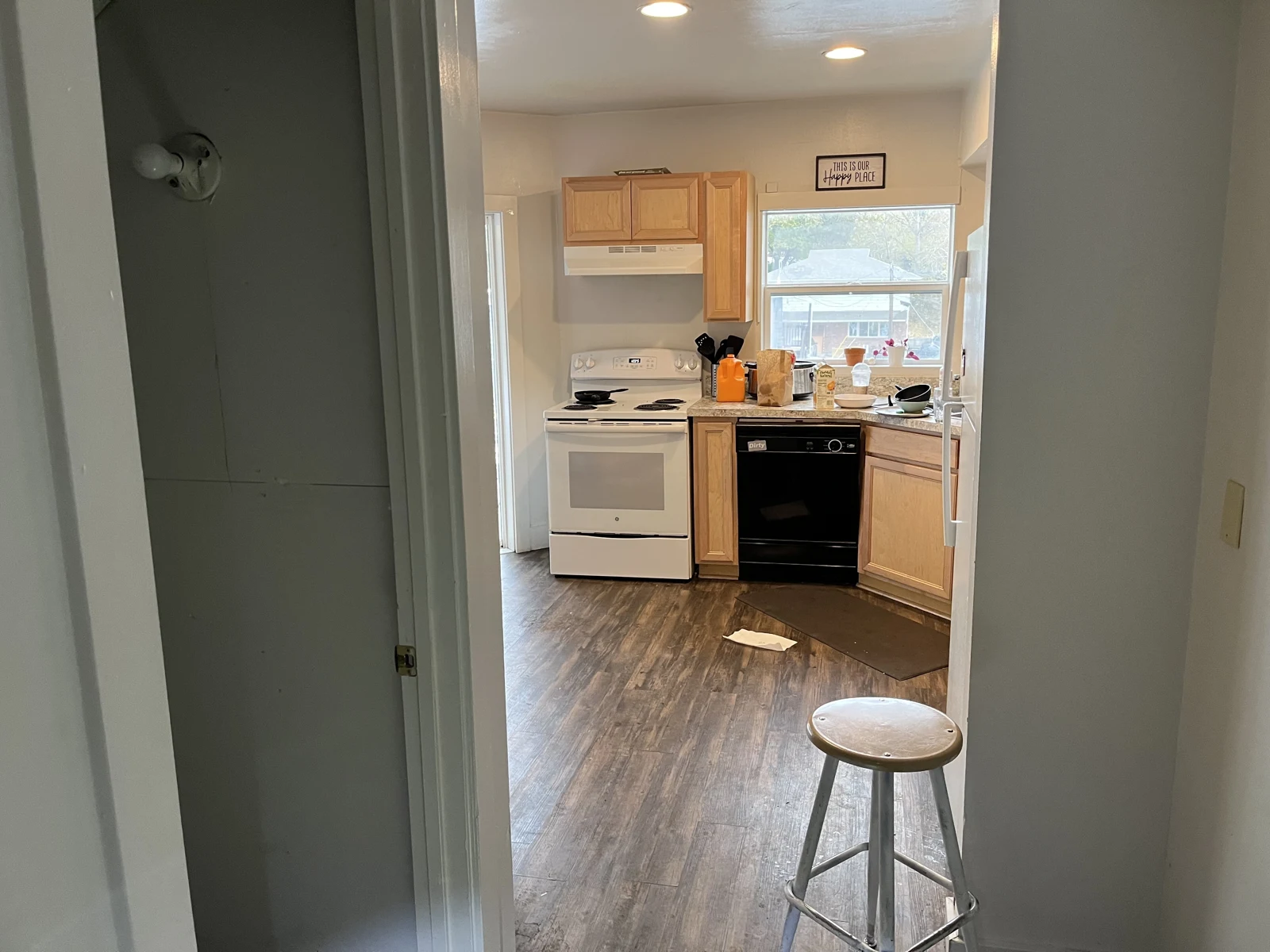 Hallway view into a kitchen with wooden cabinets, white stove, black dishwasher, window and a round stool.