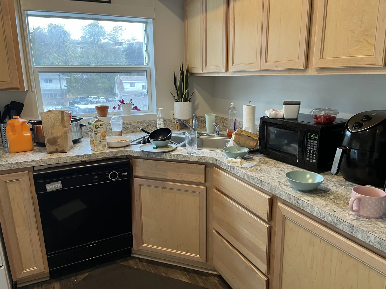 Kitchen corner counter with sink, dishes, paper towels, microwave, air fryer and wooden cabinets beneath a window.
