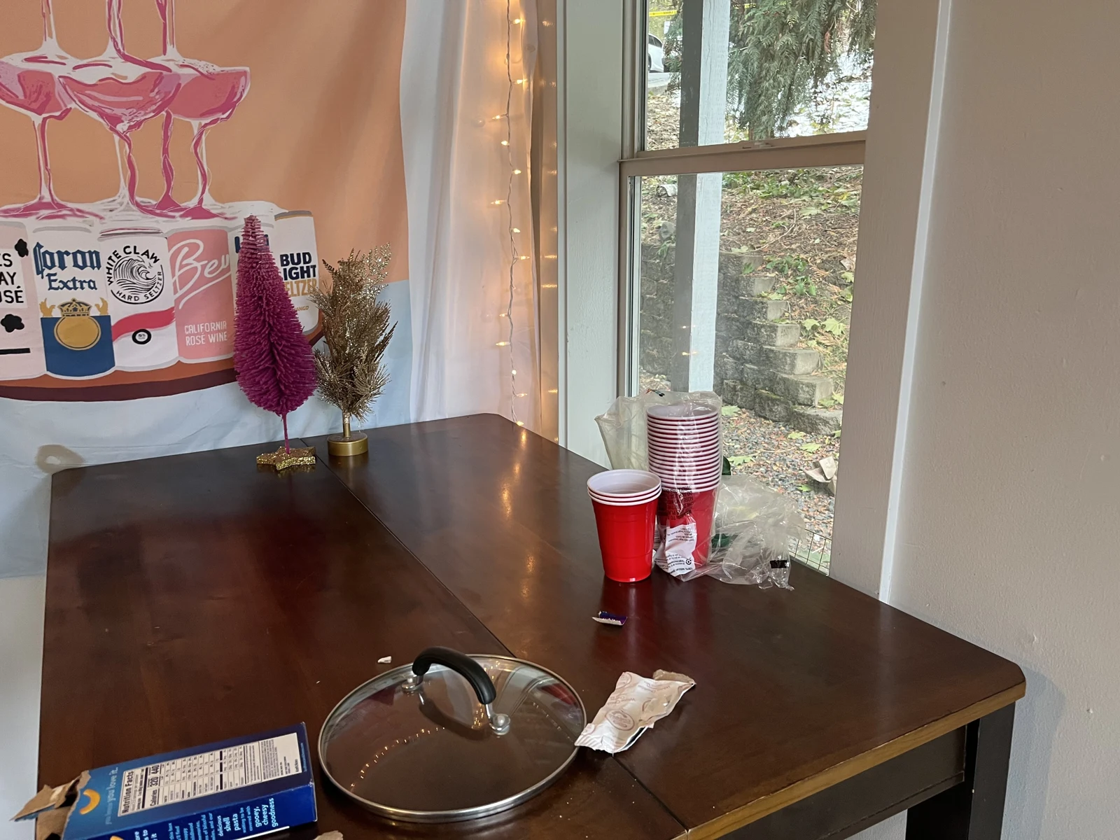 Dark table with stacked red cups, a pot lid, packaging and small decorative trees beside a window and string lights.