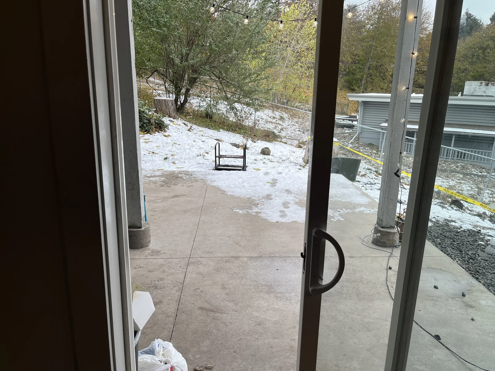 Sliding glass door looking onto a concrete patio and snowy yard, with trees, a small overturned stool and a neighboring building.