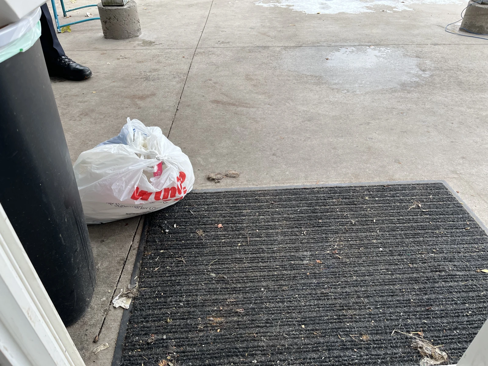 Concrete patio threshold with a dark doormat, white plastic shopping bag, trash bin edge and small debris.