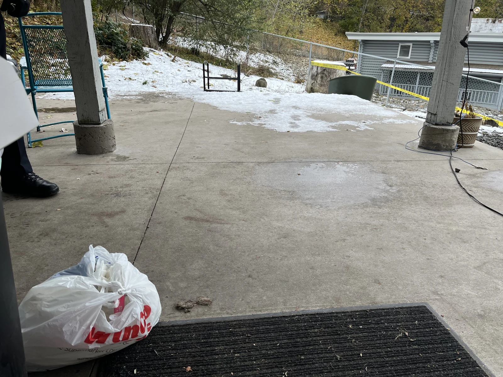 Wider concrete patio view with a dark doormat, white bag, support posts and a snowy yard beyond.