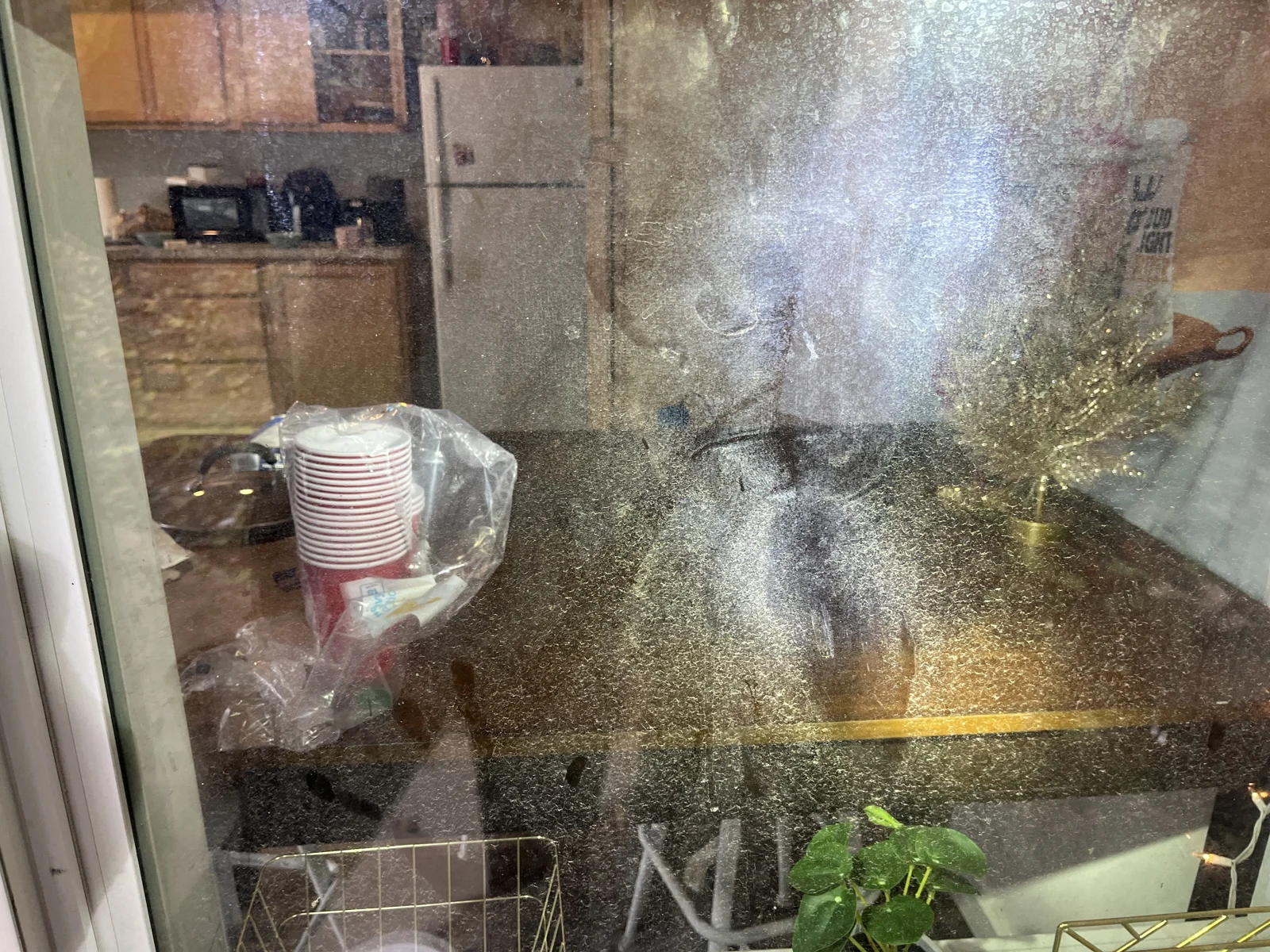 View through speckled window glass into a kitchen and dining area, with a refrigerator, microwave and stacked red cups.