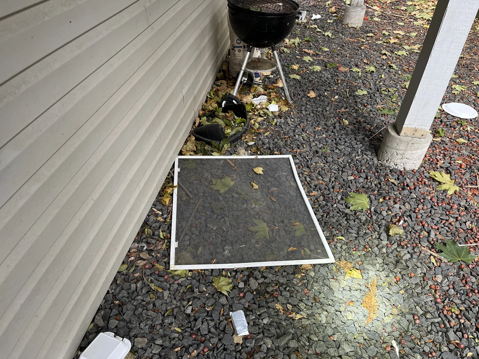 Detached window screen on gravel beside gray siding, with a small black charcoal grill, leaves and discarded packaging.