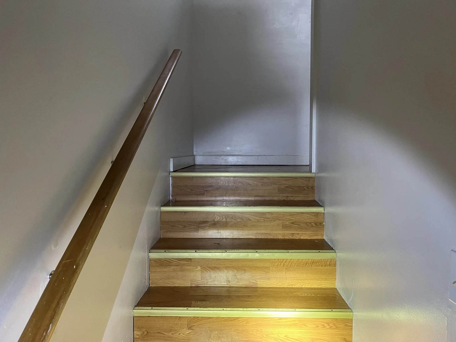 Wooden stair flight between pale walls with a wooden handrail, illuminated by a flashlight.